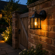 Dulwich outdoor wall light mounted on a garden brick wall beside a wooden gate at dusk