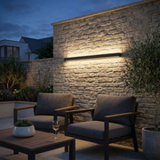 Ealing Long IP65 Linear Outdoor Wall Light illuminated on a stone garden wall at dusk beside a patio seating area