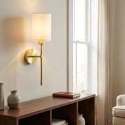 Slender brass wall light with white fabric shade above a console table, warm glow