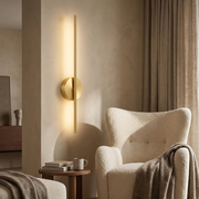 Gold vertical linear wall light glowing beside a reading chair