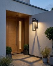 Black geometric outdoor wall light glowing beside a front door at dusk