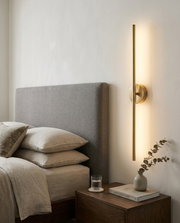 Gold rotatable linear wall light glowing on a bedside wall in a warm bedroom