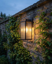 Black solar outdoor wall lantern glowing on a garden boundary wall at dusk