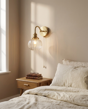 Brass plug-in wall light glowing softly beside a bed with warm bedside ambience