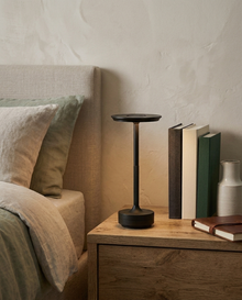 Black cordless metal table lamp glowing on a wooden bedside table