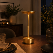Gold Pembroke cordless lamp lit on a dining table in a warm evening setting